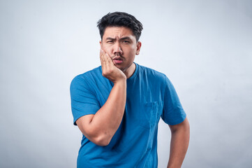 An Asian man wearing a blue t-shirt against a white background holds his cheek with a pained...