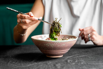 A close-up of a person using chopsticks to pick up a leafy vegetable from a bowl, set against a...