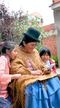 vertical aymara cholita grandmother in pollera looking at her cell phone with her two granddaughters in the backyard of her house in the city of la paz bolivia - communication concept