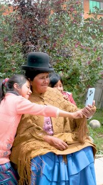 vertical aymara cholita grandmother in pollera taking a selfie with her cell phone with her two granddaughters in the courtyard of her house in the city of la paz bolivia - communication concept