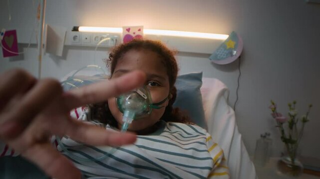 POV shot of cheerful little Black girl lying on hospital bed with oxygen mask on face, smiling at camera and showing peace sign while receiving medical aid in modern clinic