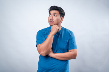 An Asian man in a blue t-shirt stands against a plain white background with his arms crossed and...
