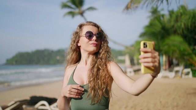 Attractive woman takes selfies on tropical beach, wavy hair, sunglasses. Smiles, poses for smartphone camera, records moments beside sea, relaxes on vacation. Influencer creates content on sunny day.