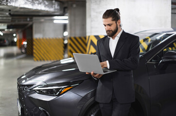 Caucasian businessman in suit using laptop near car in parking garage, indicating modern work...