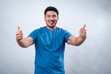 Fototapeta premium A cheerful Asian man wearing a blue t-shirt gives a thumbs-up gesture while smiling, standing against a clean white background. His positive expression and body language convey confidence and approval
