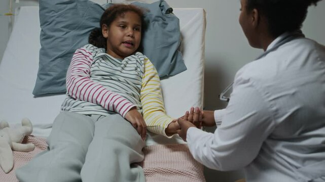 High angle slowmo of nice Black female doctor in lab coat checking pulse of smiling little Black girl lying on hospital bed, talking with care and creating positive atmosphere for recovery