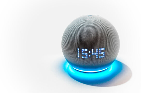 Primer plano de un dispositivo Amazon Echo Dot (4&ordf; generaci&oacute;n) en color gris claro con luz azul activada y mostrando la hora sobre un fondo blanco.