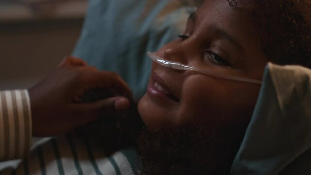 Close up shot of unrecognizable caring mother gently adjusting pyjamas and braids of smiling little Black girl lying in hospital bed with nasal cannula showing her support