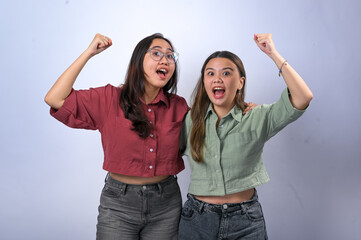 Two young Asian women with long hair wear casual crop shirts, one red and one green, with dark jeans. They cheer with raised fists while standing close together against a white background.