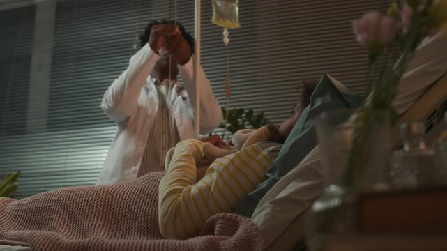 Young Black female doctor in lab coat setting dropper for little Black girl lying in hospital bed in evening hugging favorite toy, then talking to her gently and leaving ward