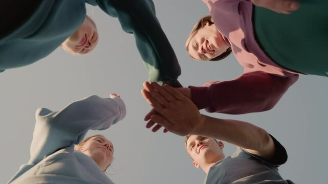 Upward view of people in hoodies placing their hands together as team members with sunlight creating a glowing effect around them, capturing team spirit and unity as they smile in outdoor setting