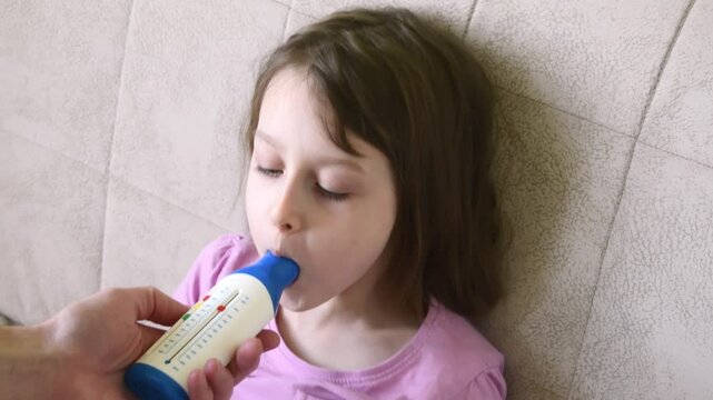 A girl uses a peak flow meter to measure lung function while sitting on a couch. An adult holds the device, assisting in respiratory health monitoring.