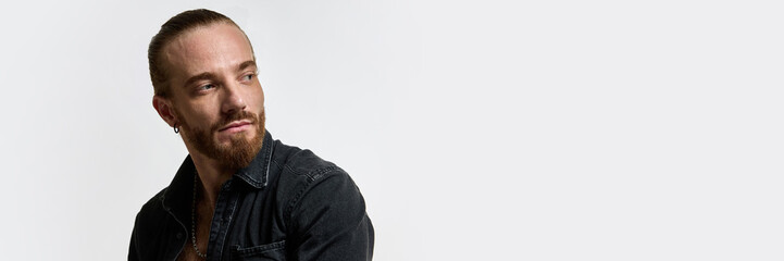 Obraz premium Man with tied-back hair and beard looking sideways, wearing dark denim shirt, expressing confidence against white background. Concept of male beauty, skin care, fashion. Banner. Copy space for text