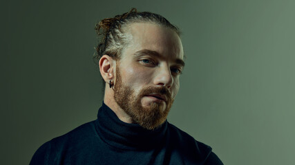 Obraz premium Portrait of handsome man with tied up hair and beard, wearing black turtleneck, posing with confidence against green studio background. Concept of male beauty, fashion. Copy space