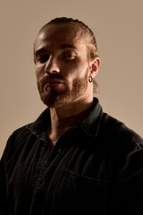 Portrait of handsome bearded man with tied up hair, in black shirt design with confidence against warm toned background. Concept of male beauty, skin care, fashion, wellness
