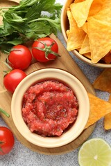 Spicy salsa sauce in bowl, nachos and ingredients on light grey table, flat lay