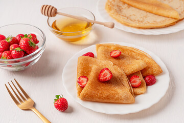 Fried homemade folded crepe french dessert or sweet blini tasty snack made from batter served on plate with sliced juicy red strawberries on white wooden table with honey, dipper and fork for lunch