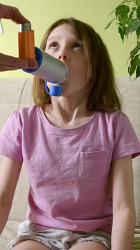 Girl sitting on couch using inhaler with spacer while adult assists. Pediatric asthma treatment, respiratory therapy, controlled breathing.