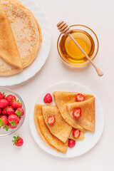 Flat lay with homemade sweet folded crepe french dessert or sweet blini tasty snack made from batter served on plate with red strawberries on white wooden table with bowl of honey, dipper and berries