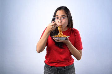 Asian woman in a red shirt and glasses enthusiastically eating instant noodles with a fork while...