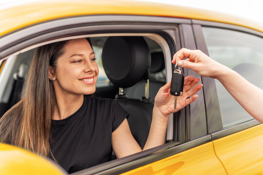 A smiling woman sitting in a yellow car receives a set of keys from another person, symbolizing a car purchase, rental, or ownership transfer.
