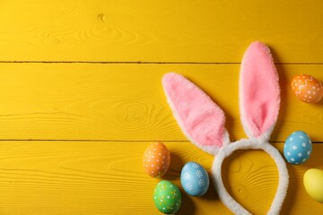 Beautiful decorated Easter eggs and bunny ears headband on yellow wooden table, flat lay. Space for text