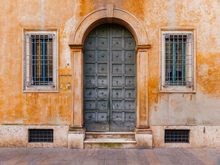 A vintage, arched, wooden door and two windows on an ocher colored wall by the sidewalk. Travel to...