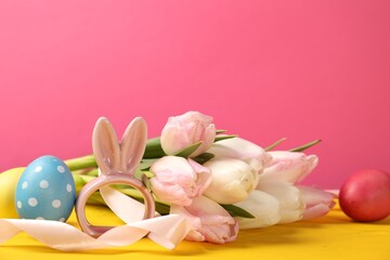 Easter eggs, beautiful tulips and bunny figure on yellow wooden table against pink background