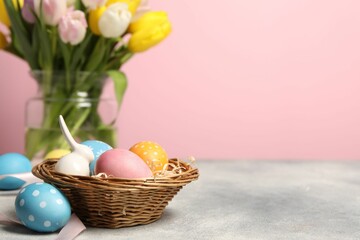 Easter eggs in wicker basket, bunny figure and beautiful tulips on grey table against pink background, closeup. Space for text