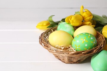 Easter eggs in wicker basket and beautiful tulips on white wooden table, closeup. Space for text