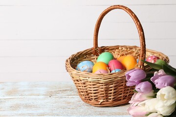 Easter eggs in wicker basket and beautiful tulips on wooden table, space for text