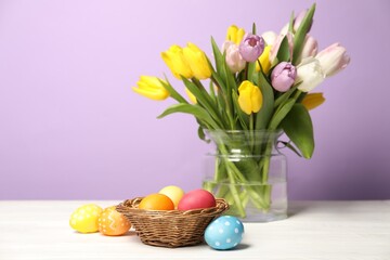 Easter eggs in wicker basket and beautiful tulips on white wooden table against lilac background