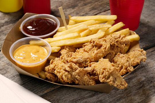 batter fried chicken tender strips with French fries and dipping sauces in paper food basket on rustic wood surface