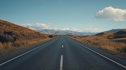 Naklejka premium Endless asphalt road stretches through golden grassy hills towards distant mountains, symbolizing journeys and exploration. Perfect for travel, adventure, and landscapes.