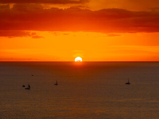 The image shows a sunset over calm ocean waters in Oahu, Hawaii, with sailboats scattered across the sea and an orange yellow glow in the sky.