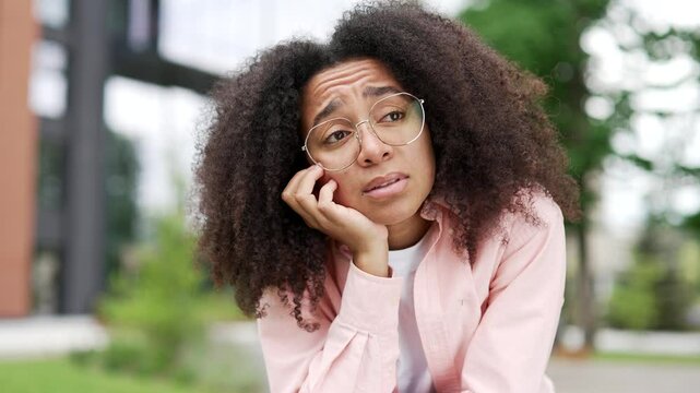 Sad young african american female feels lonely and bored sitting on a bench on street near university building. Upset black woman is depressed and in a bad mood, suffering from mental pain. Close up