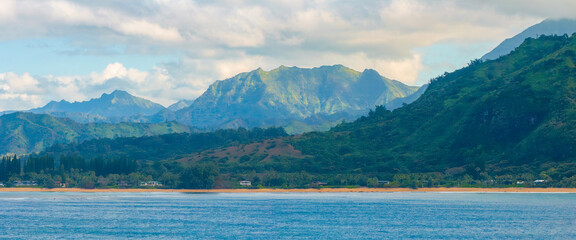 Kauai Island's Napali cliffs rise above lush greenery and a sandy beach, bordered by calm blue ocean and scattered small structures under a cloudy sky.