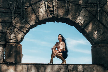 A woman with long black hair and glasses is squatting on a stone arch. She is wearing a light pink blouse and a black skirt