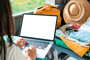 Happy traveler Asian woman planning vacation trip with laptop and packing luggage at home. Getting ready for summer holiday trip. Blank screen for copy space