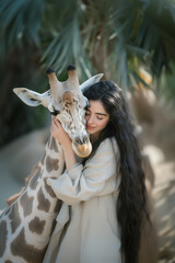 Woman hugs giraffe. Long-haired female model in linen dress cuddles cute baby giraffe at petting zoo