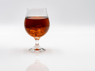 A crystal glass filled with a dark amber beverage sits on a smooth, reflective white surface. Ice cubes float in the drink, adding a refreshing touch to the presentation.