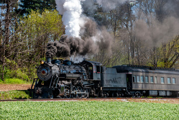 A vintage steam locomotive chugs along a green field, releasing thick smoke into the clear sky. This picturesque journey showcases a blend of history and nature, perfect for train enthusiasts.