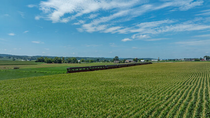 A vintage train travels through expansive green fields under a bright blue sky. Farms and distant hills provide a picturesque backdrop, showcasing the beauty of rural life.