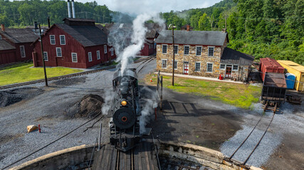 A vintage steam locomotive steams into a train yard during the afternoon. Nearby, historic buildings surround the area, showcasing a charming rural landscape.