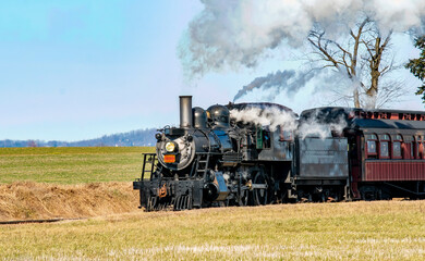 Obraz premium A vintage steam locomotive chugs along rural tracks, releasing puffs of white smoke against a bright blue sky. The train is moving steadily through expansive green fields.