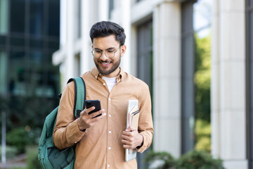 Young student with backpack on shoulder using app on smartphone. Man walking outside university...