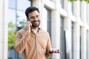 Young smiling man talking on the phone while walking outside the office building. Company employee in casual clothes, satisfied and successful programmer student.