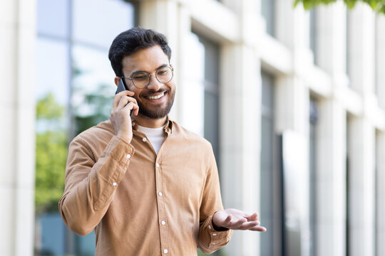 Young smiling man talking on the phone while walking outside the office building. Company employee in casual clothes, satisfied and successful programmer student.