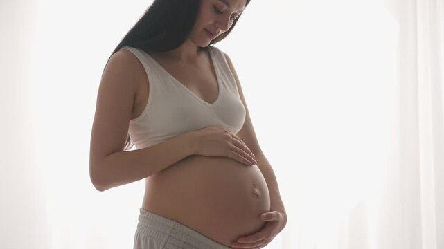 Young happy Asian pregnant woman posing in studio with white curtains in background