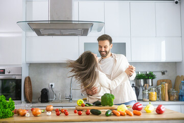 A couple shares a playful dance moment in a well-decorated kitchen, surrounded by colorful vegetables, enhancing their relationship.